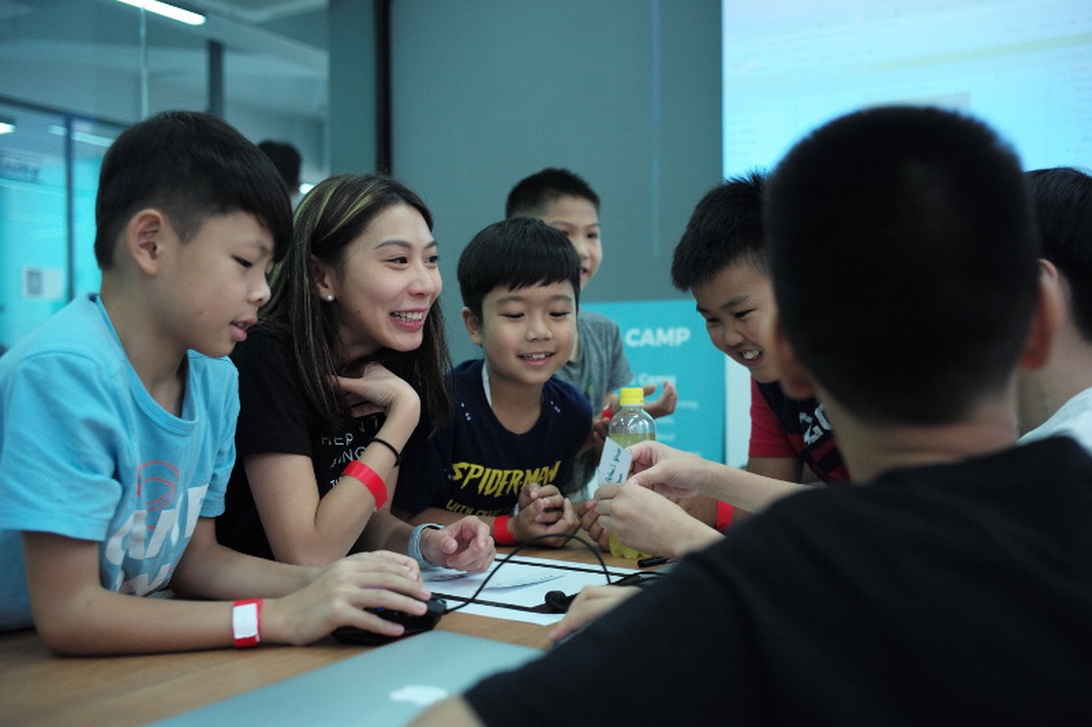 A group of children gathered around a table with what appears to be technology components or a project. An adult facilitator is engaging with them in what looks like a structured educational environment, possibly a tech camp or after-school program. The children wear colored wristbands suggesting an organized program. This represents community technology education for young learners.