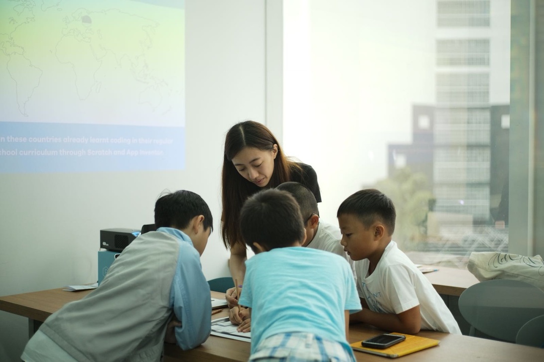A small group of children are gathered around a table with an adult instructor or facilitator leaning in to guide their work. A projector screen in the background displays what appears to be educational content, suggesting this is a structured technology education session. The setting appears to be a classroom or community center dedicated to technology learning.
